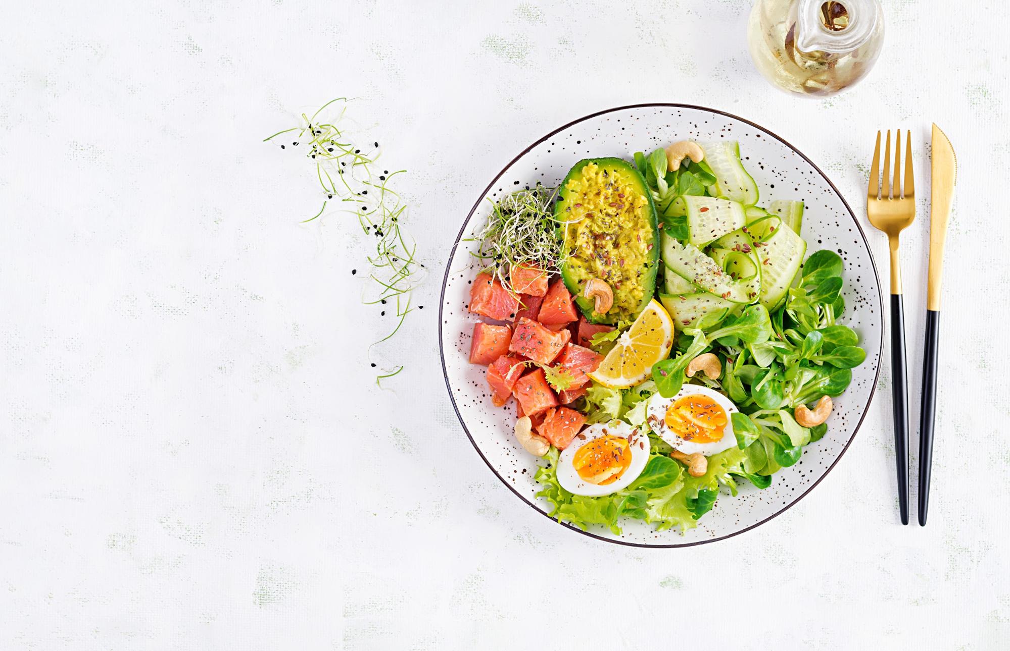 A plate of fruits, vegetables and eggs with gold knife and fork beside it on a white surface