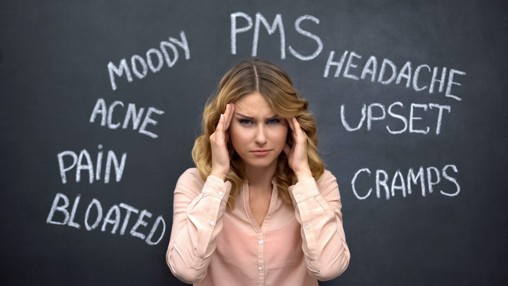 Woman holding her head in pain while symptoms of hormonal imbalance in women are written on a blackboard behind her