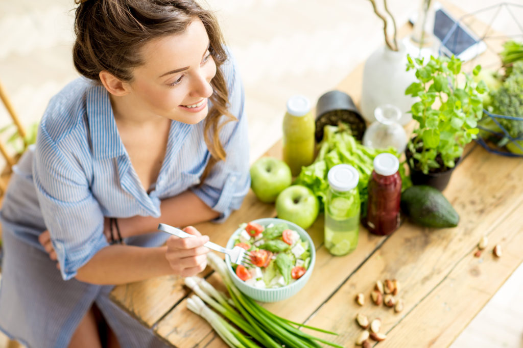 Smiling woman leaning on a wooden table with a bowl of salad and fresh ingredients in front of her